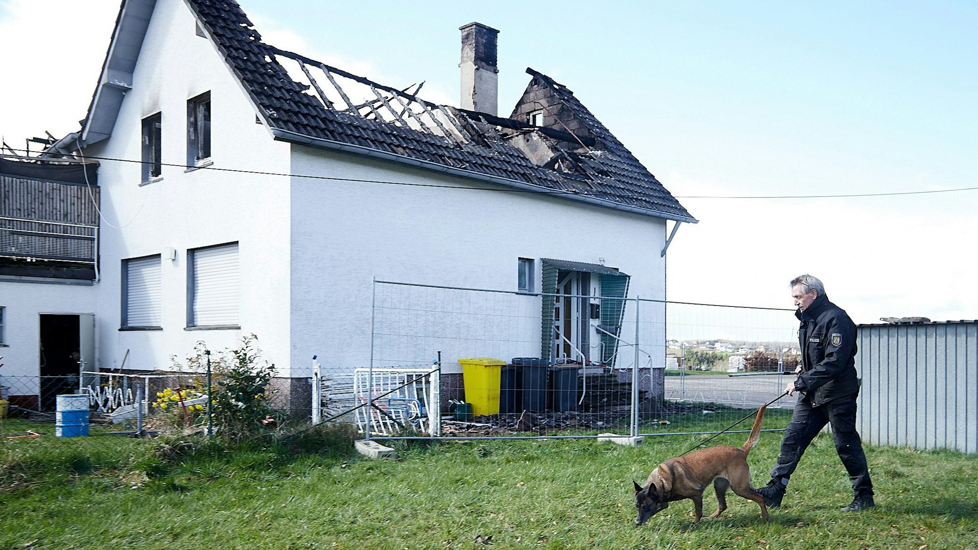 Vor einem ausgebrannten Haus geht ein Polizist mit der Brandspürhündin Saphira über eine Wiese.