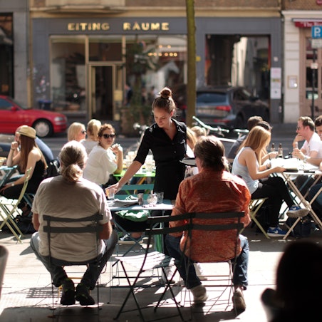 Menschen sitzen in einer Straße in der Außengastronomie an Tischen. Eine Kellnerin bedient sie.