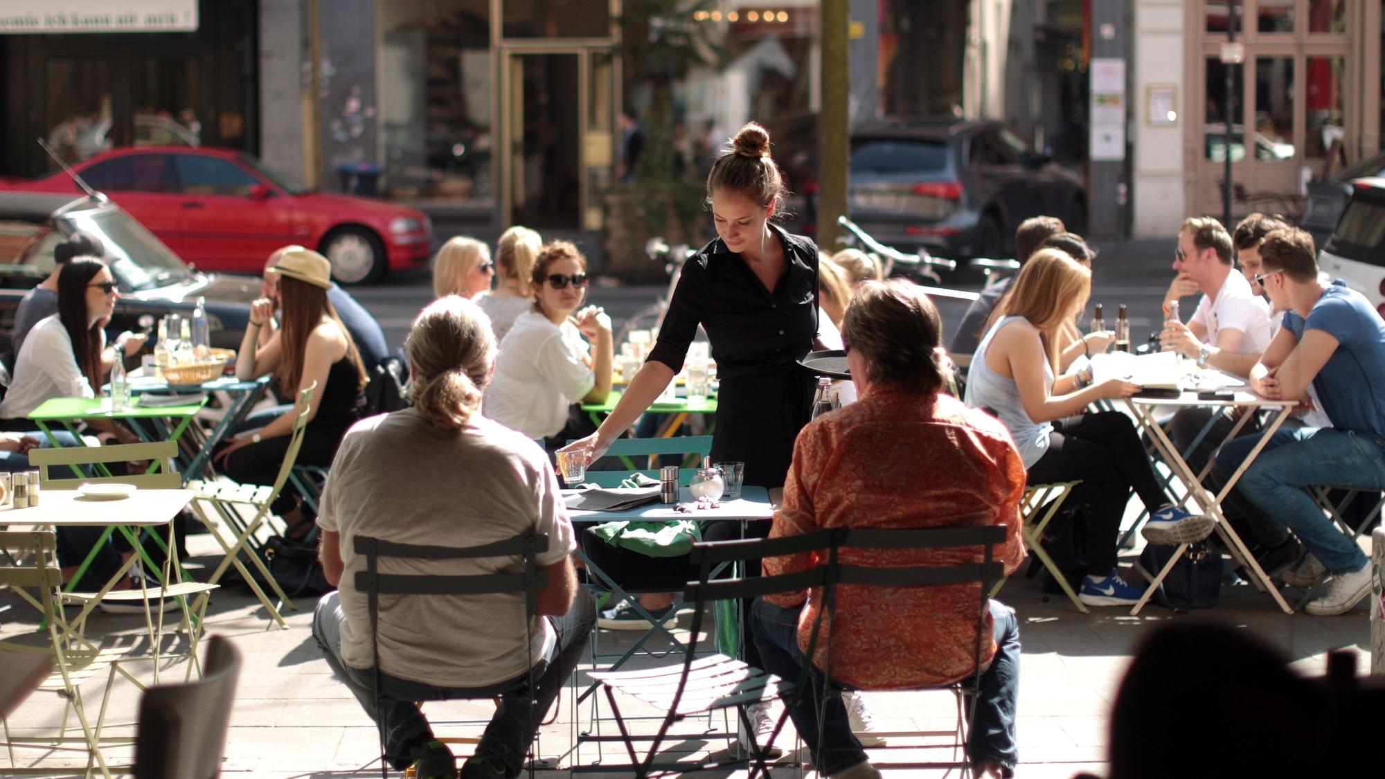 Menschen sitzen in einer Straße in der Außengastronomie an Tischen. Eine Kellnerin bedient sie.