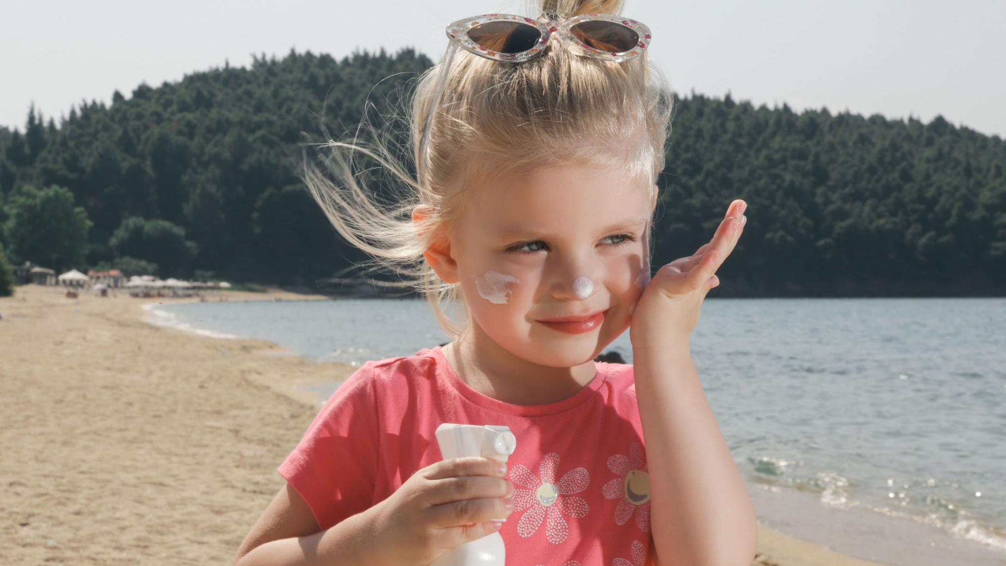 Kleines Mädchen steht an einem See und cremt sich mit Sonnenmilch ein.