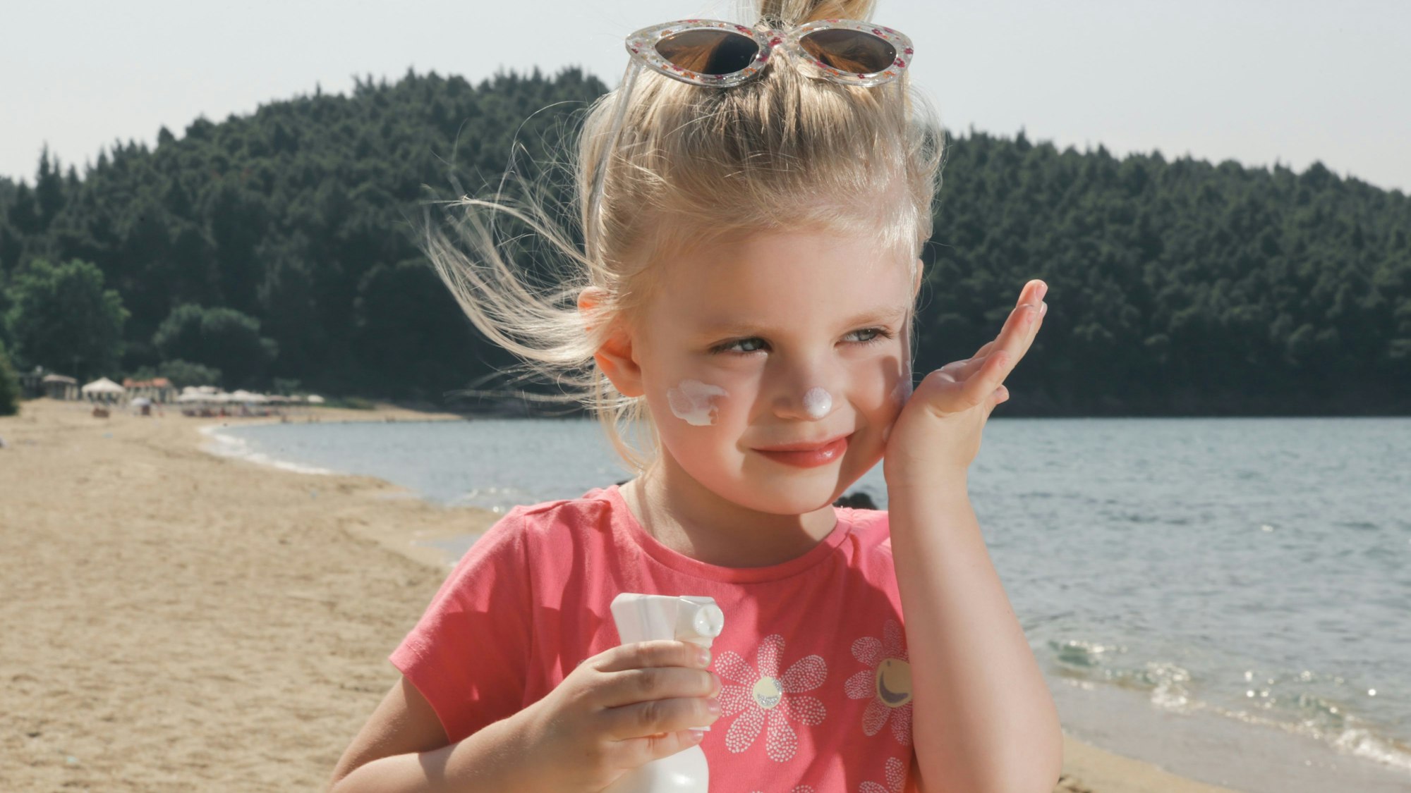 Kleines Mädchen steht an einem See und cremt sich mit Sonnenmilch ein.