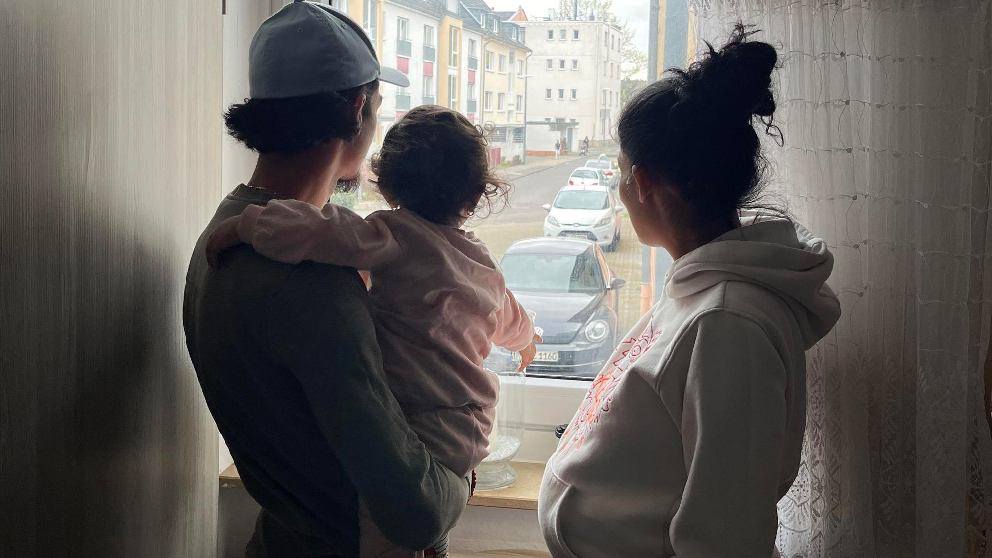 Silhouette einer dreiköpfigen Familie: Vater mit Tochter auf dem Arm, Mutter rechts, gucken aus dem Fenster