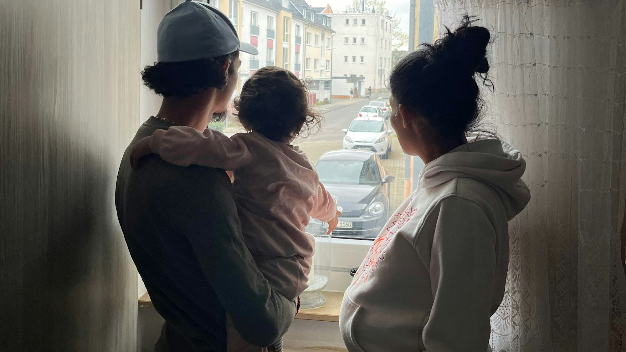 Silhouette einer dreiköpfigen Familie: Vater mit Tochter auf dem Arm, Mutter rechts, gucken aus dem Fenster