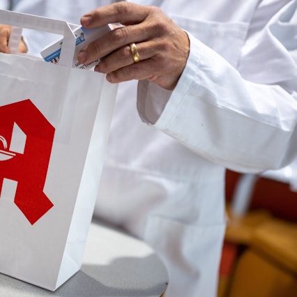 Ein Apotheker legt in einer Apotheke eine Packung mit Tabletten in eine Papiertasche mit Apotheken-Logo.