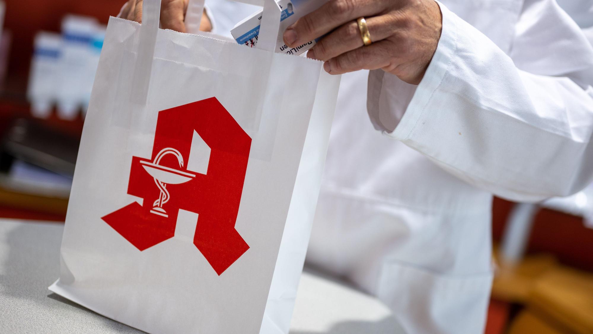 Ein Apotheker legt in einer Apotheke eine Packung mit Tabletten in eine Papiertasche mit Apotheken-Logo.