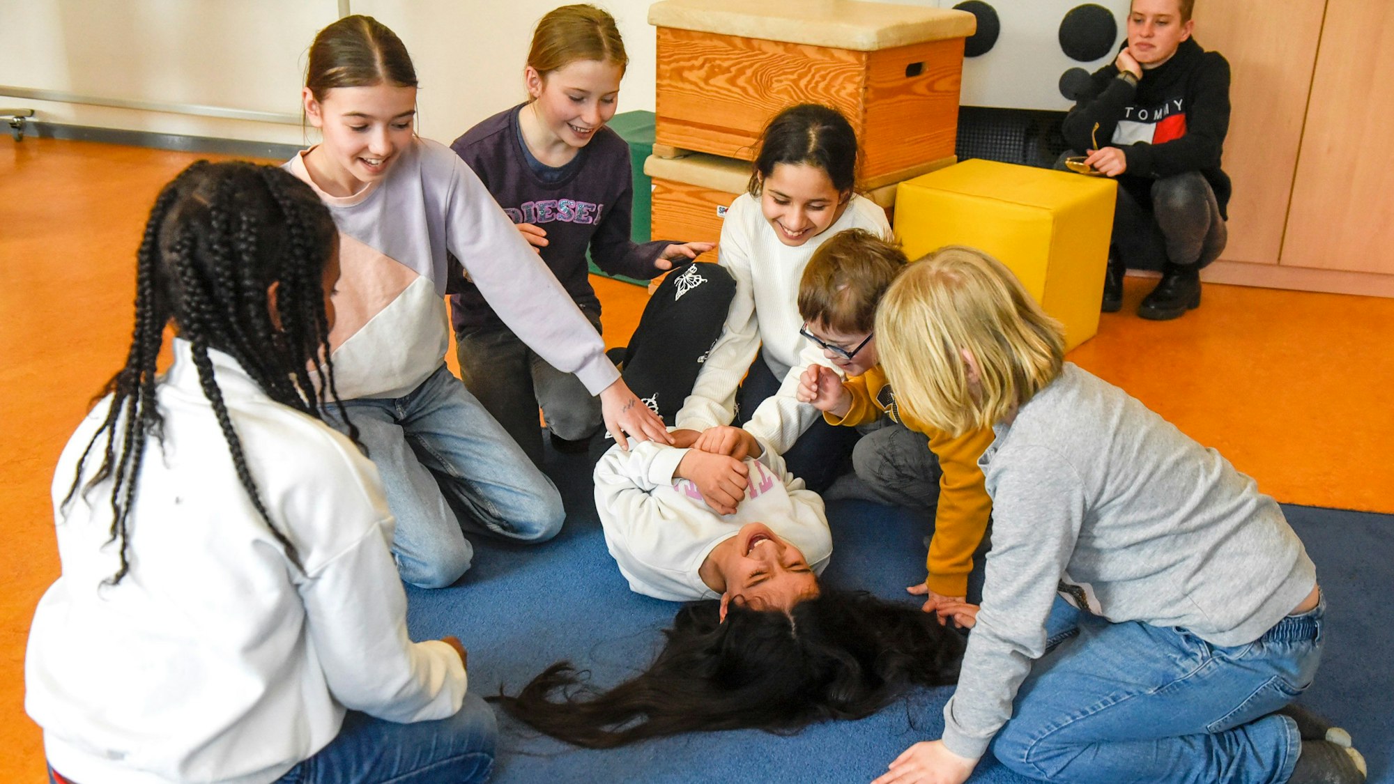 Sechs Kinder sitzen auf dem Boden, in ihrer Mitte liegt ein Mädchen, das von einigen ausgekitzelt wird.