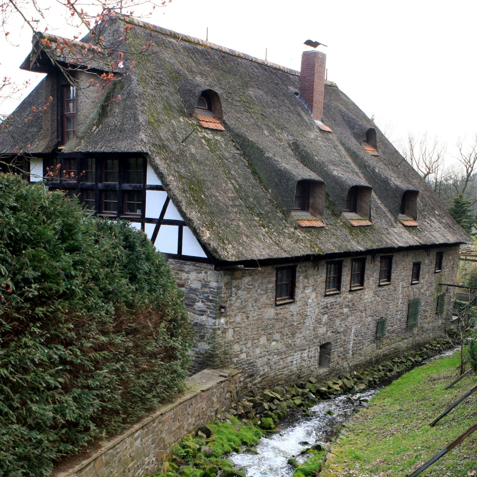 An einem Bach steht ein reetgedecktes Haus.