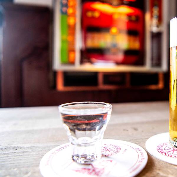Im Vordergrund stehen ein Kölsch und ein Schnaps, im Hintergrund ist ein Spielautomat zu sehen.