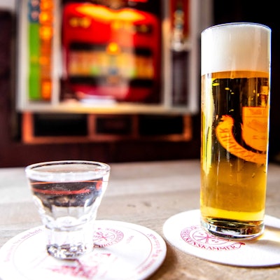 Im Vordergrund stehen ein Kölsch und ein Schnaps, im Hintergrund ist ein Spielautomat zu sehen.