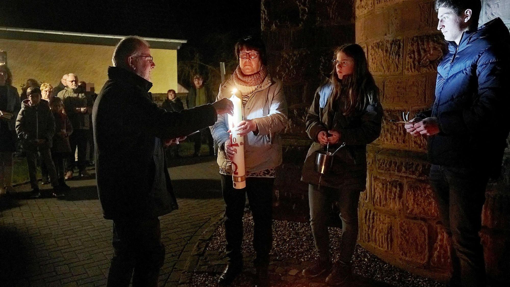 Vier Personen stehen um eine Osterkerze, ein Mann entzündet sie. Im Hintergrund stehen weitere Gläubige und beobachten die Szene.