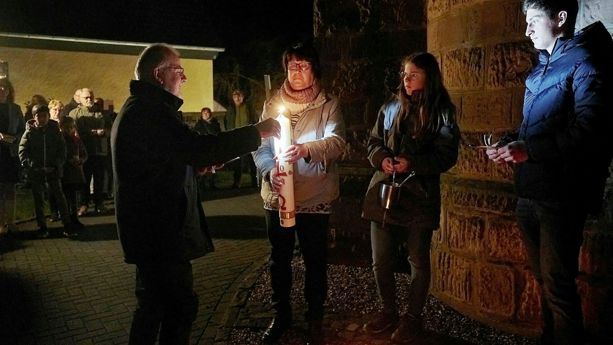 Vier Personen stehen um eine Osterkerze, ein Mann entzündet sie. Im Hintergrund stehen weitere Gläubige und beobachten die Szene.