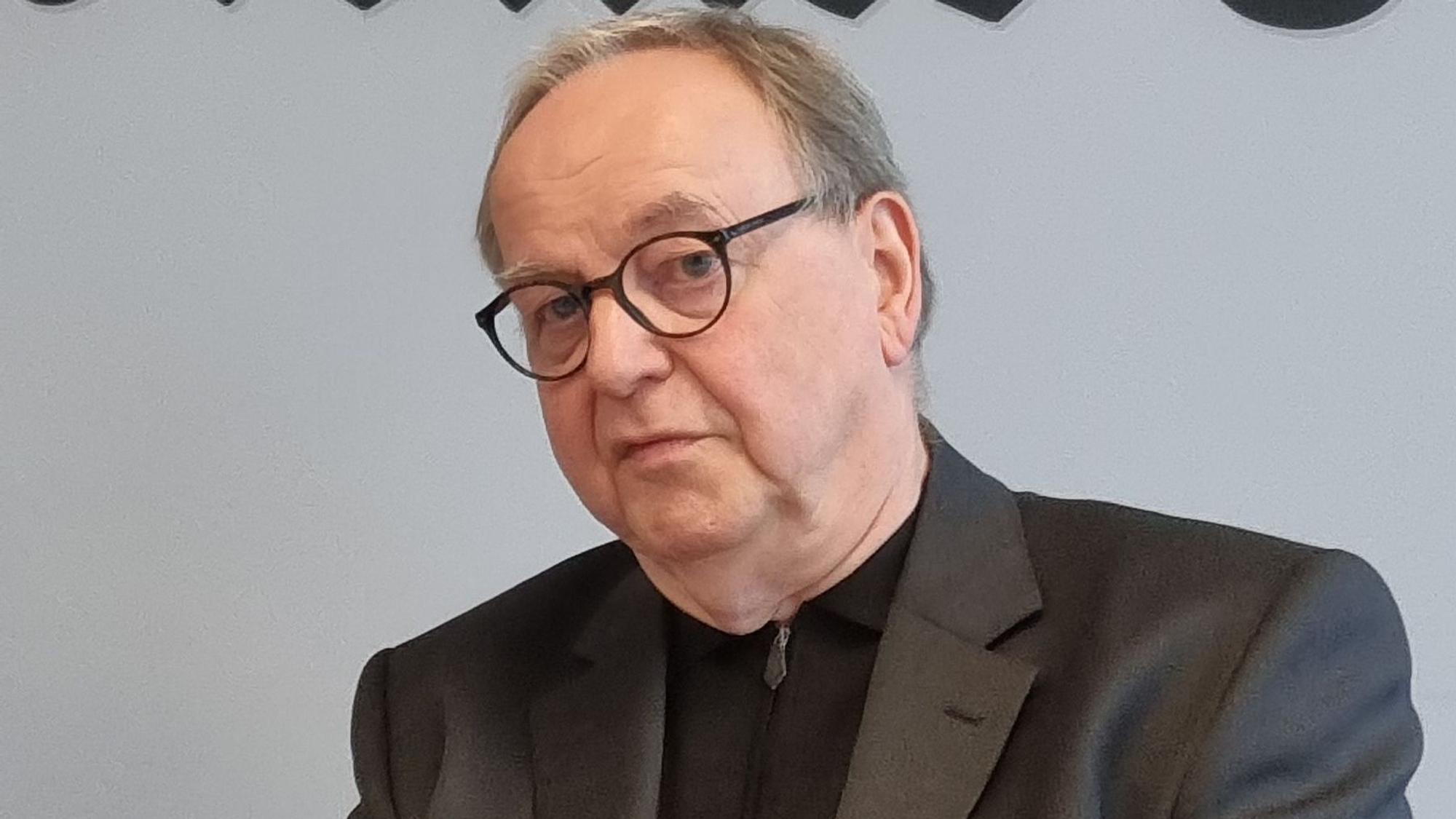 Das Bild zeigt einen sitzenden Mann vor einer Wand.