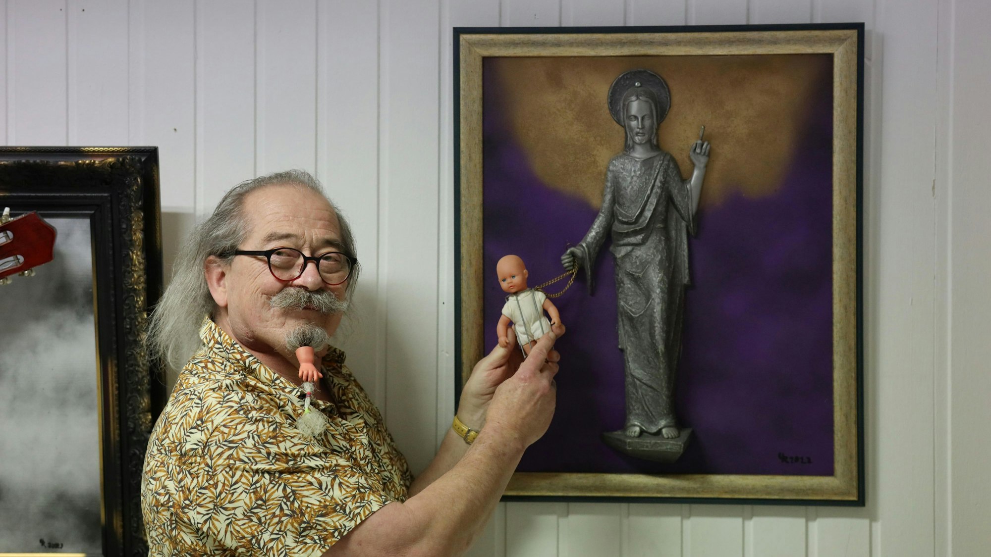 Uwe Rhiem hält eine Puppe vor ein Bild mit einer Christusfigur.