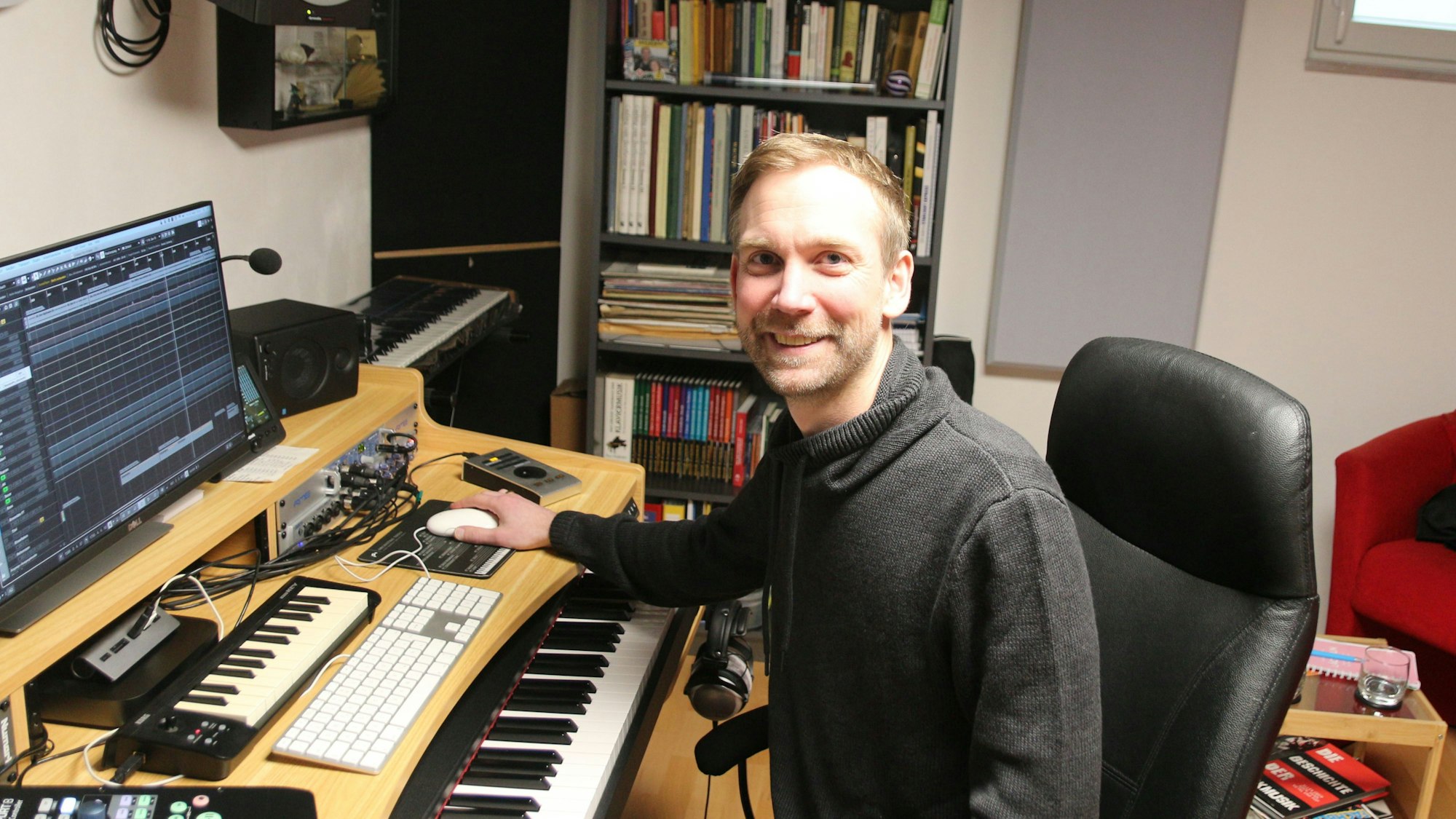 Der Musiker sitzt in seinem Tonstudio und lächelt in die Kamera.