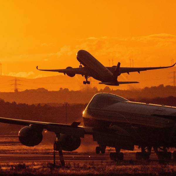 Ein Flugzeug steht bei Sonnenuntergang auf einem Rollfeld. Im Hintergrund hebt eine anderes Flugzeug ab