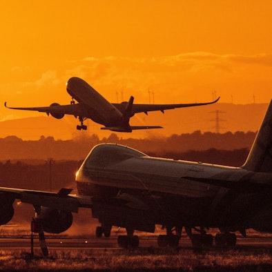 Ein Flugzeug steht bei Sonnenuntergang auf einem Rollfeld. Im Hintergrund hebt eine anderes Flugzeug ab