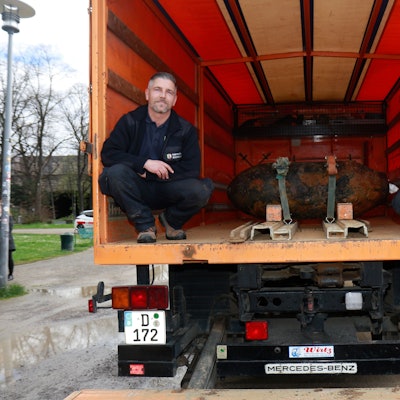 Zwei Männer in Dienstkleidung im Innenraum eines Lastwagens, in ihrer Mitte ist eine entschärfte Bombe festgeschnallt