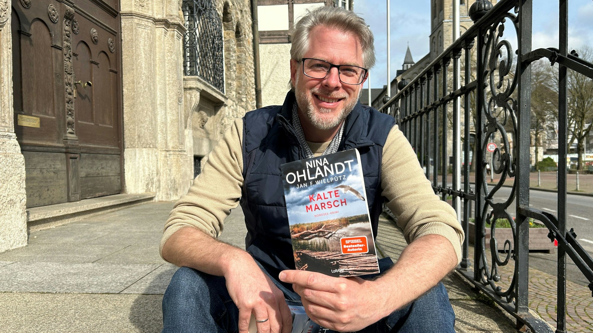 Autor Jan Wielpütz sitzt auf der Rathaustreppe in Bergisch Gladbach und zeigt seinen neuen Krimi „Kalte Marsch“