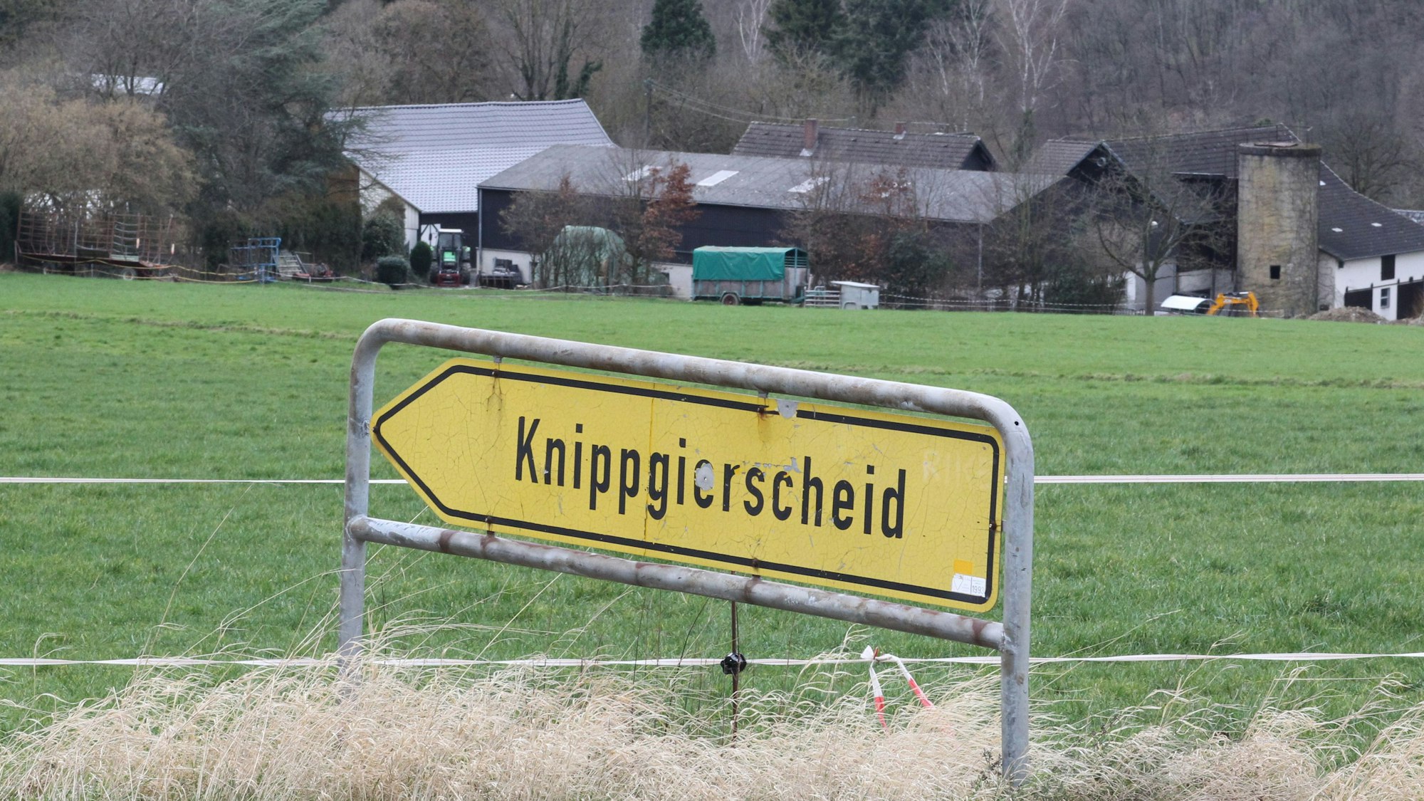 Ein gelbes Hinweisschild mit der Aufschrift Knippgierscheid vor einer Wiese und einem Bauernhof.