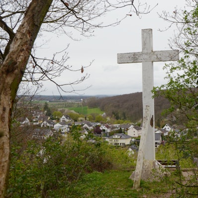 Das Kreuz steht am Waldrand, davor ist eine Sitzbank platziert. Im Bildhintergrund das Erfttal und der Ort Kreuzweingarten.