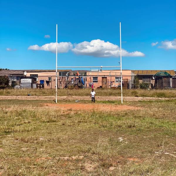Ein Kind steht vor den Stangen eines Rugby-Tors auf einem Sportplatz in Südafrika. Im Hintergrund sind die Häuser einer Township zu sehen.