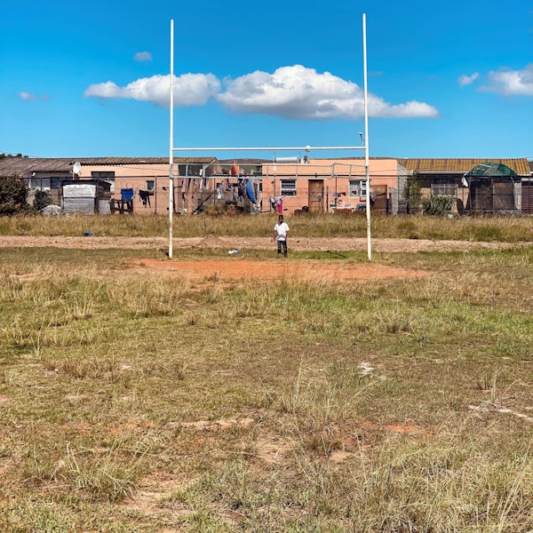 Ein Kind steht vor den Stangen eines Rugby-Tors auf einem Sportplatz in Südafrika. Im Hintergrund sind die Häuser einer Township zu sehen.