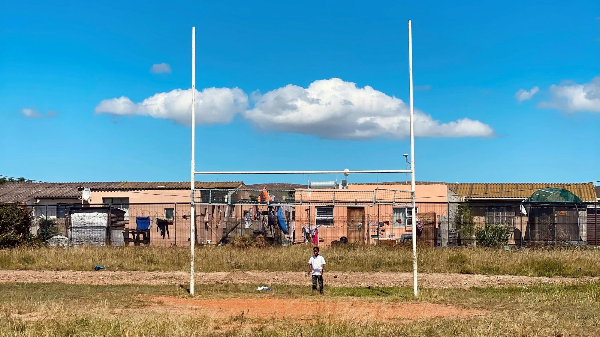 Ein Kind steht vor den Stangen eines Rugby-Tors auf einem Sportplatz in Südafrika. Im Hintergrund sind die Häuser einer Township zu sehen.