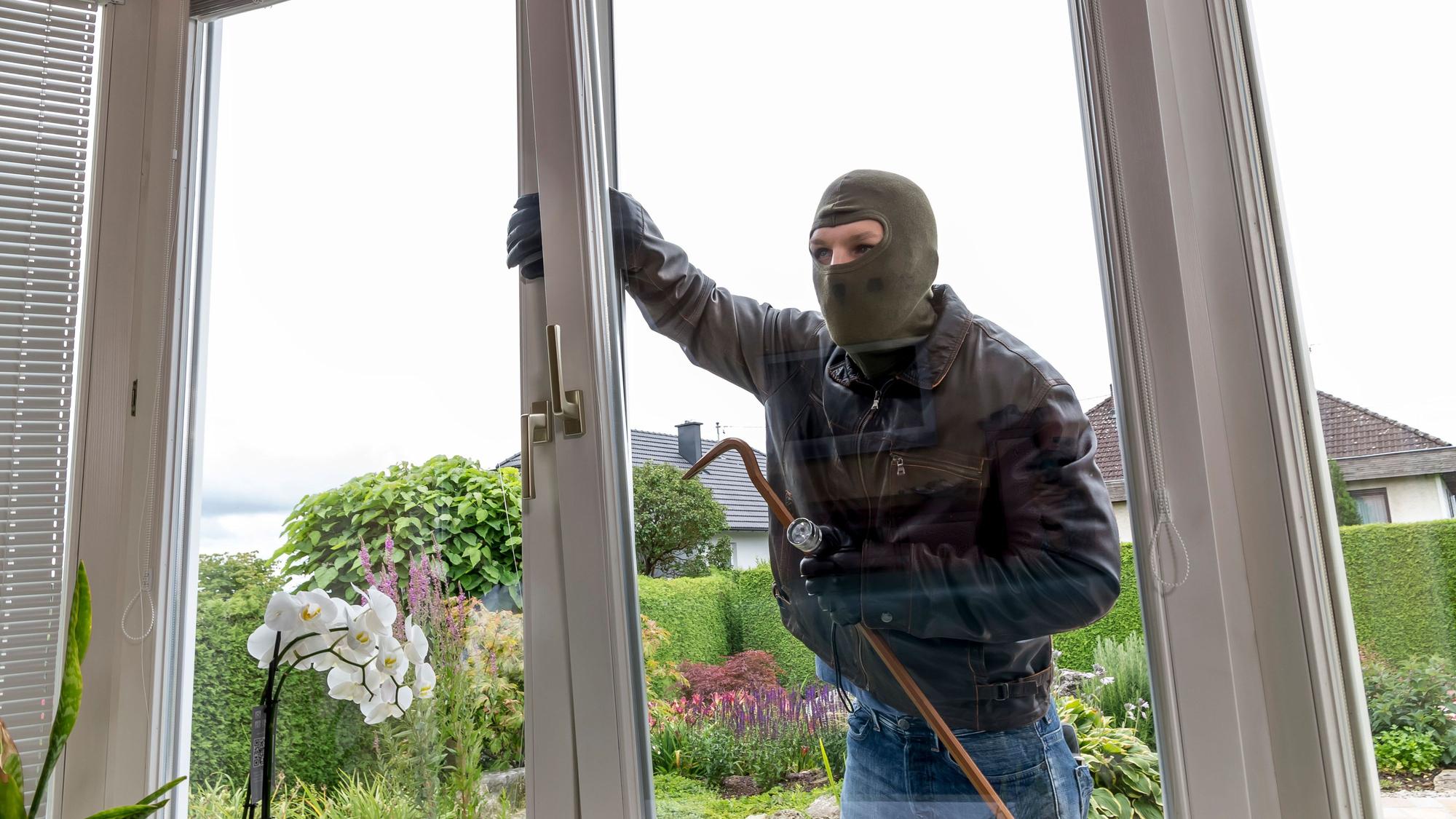 Das Bild ist ein Symbolfoto und zeigt einen maskierten Mann, der versucht, in ein Haus einzubrechen.