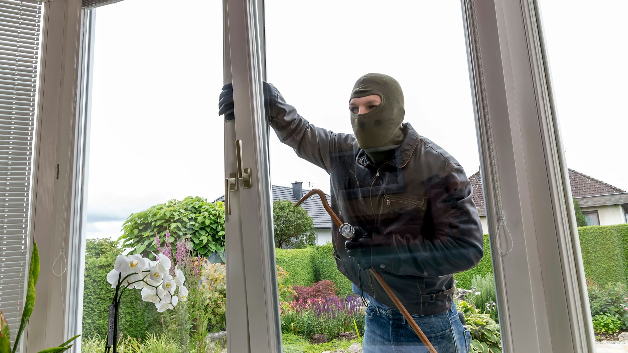 Das Bild ist ein Symbolfoto und zeigt einen maskierten Mann, der versucht, in ein Haus einzubrechen.