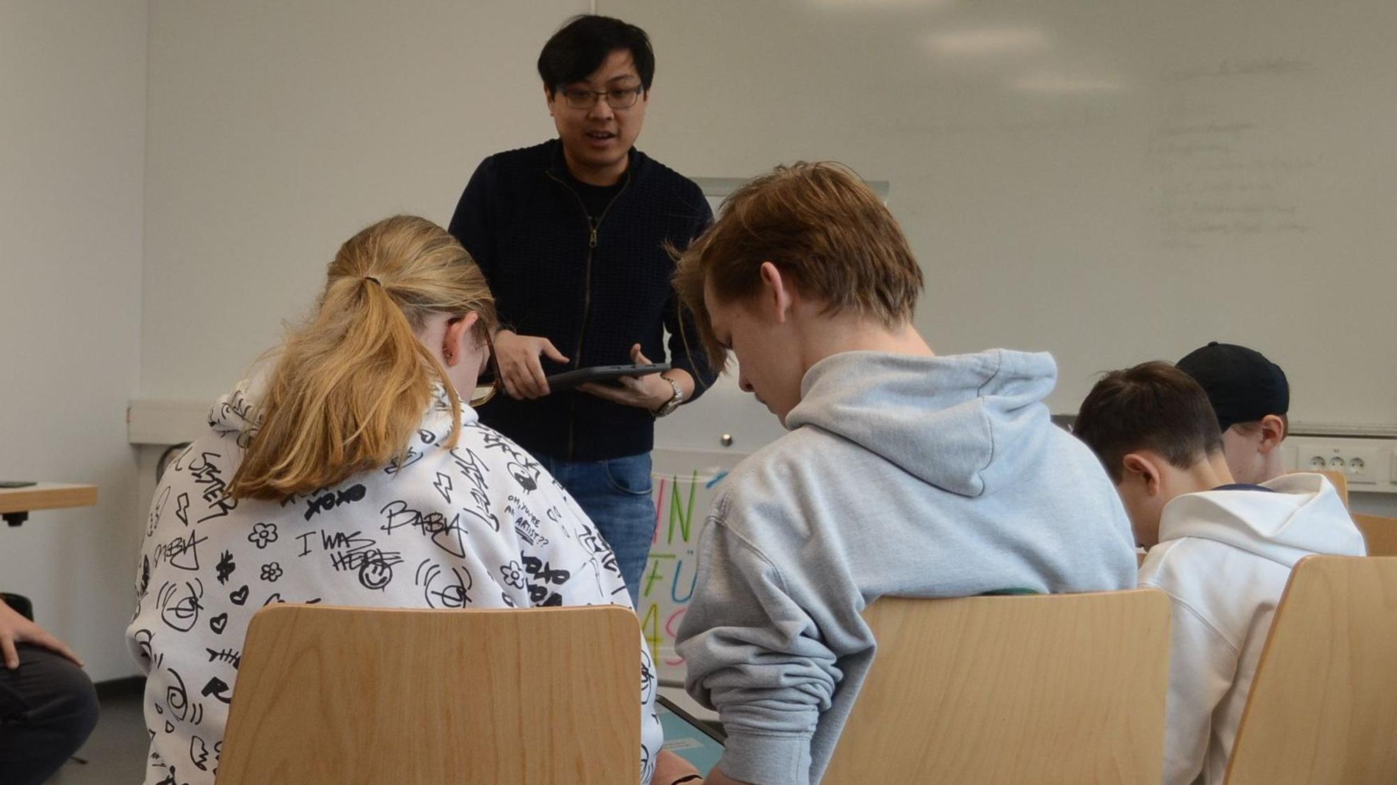 Kinder und Jugendliche beugen sich über Tablet-Computer auf ihrem Schoß. Ein Dozent steht in der Mitte.