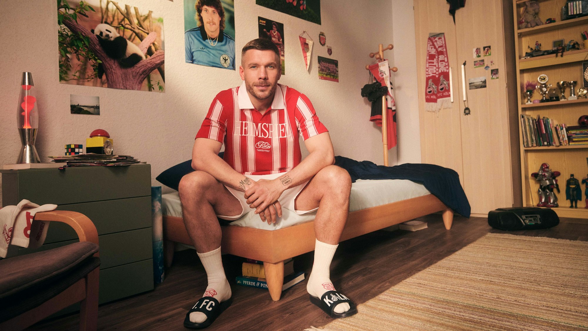 Lukas Podolski sitzt in einem Jugendzimmer auf dem Bett. Er trägt ein rot-weiß gestreiftes Poloshirt.