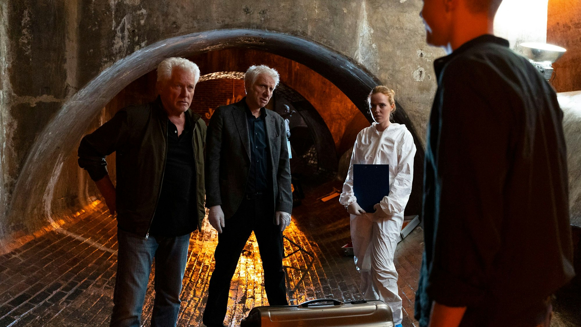 Auf dem Bild sind die Ermittler in einem Tunnel zu sehen. Kalli Hammermann ist im Vordergrund, die beiden älteren Ermittler und die Rechtsmedizinerin schauen ihn an.