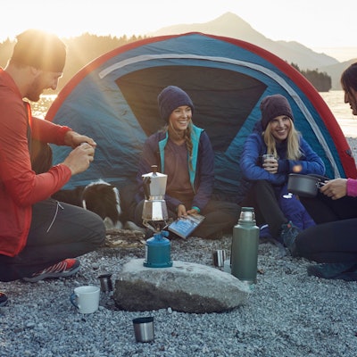 Eine Trekking-Gruppe kocht vor einem Zelt Kaffee auf einem Gaskocher.