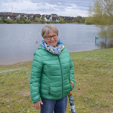 Eine Frau mit grüner Daunenjacke steht am Ufer eines Sees. Im Hintergrund sind Häuser jenseits des Sees zu sehen.