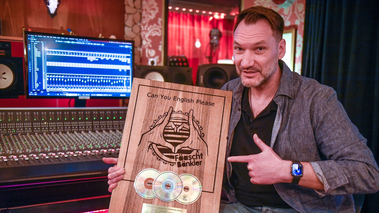 Ein Mann sitzt in einem Tonstudio vor einem Mischpult mit Bildschirm und zeigt eine Holztafel mit drei eingelassenen CDs.