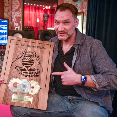 Ein Mann sitzt in einem Tonstudio vor einem Mischpult mit Bildschirm und zeigt eine Holztafel mit drei eingelassenen CDs.