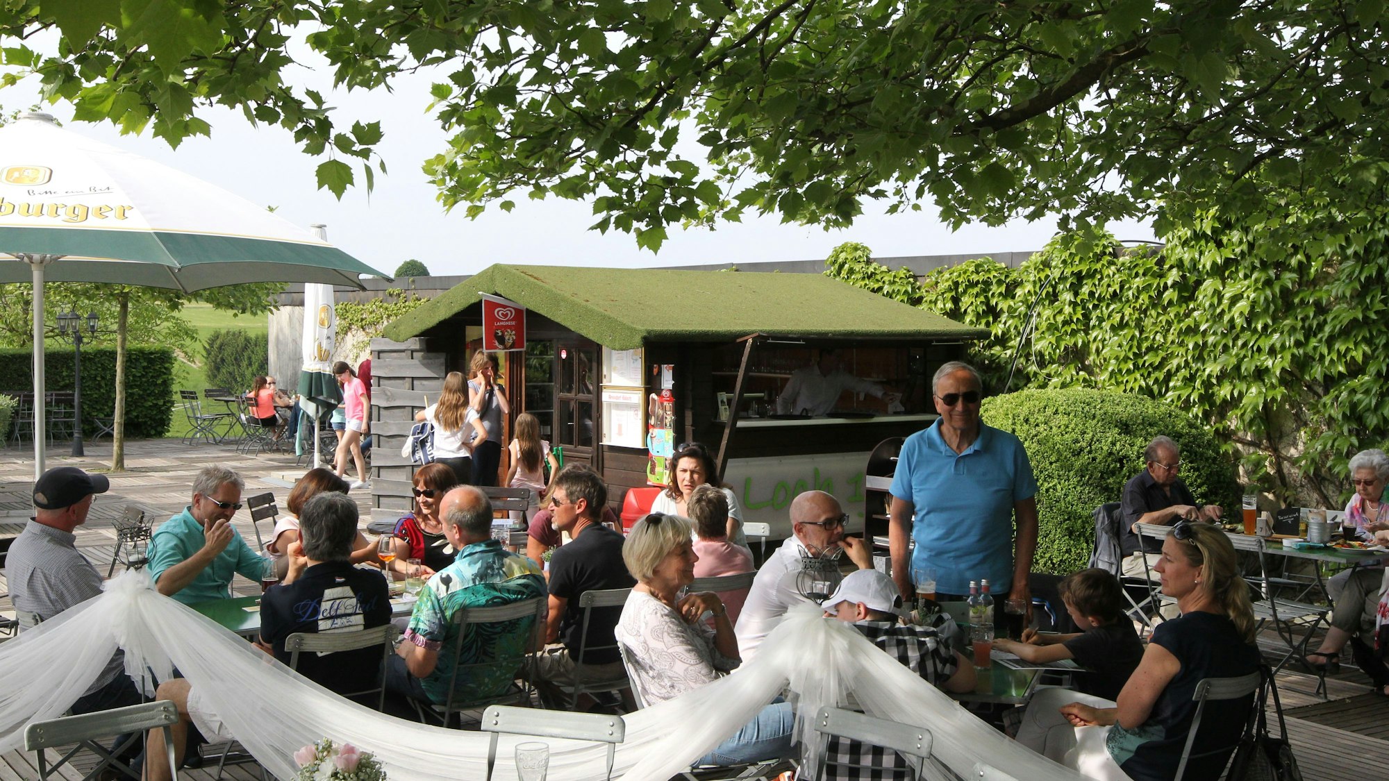 Menschen sitzen an Biertischen unter Bäumen.