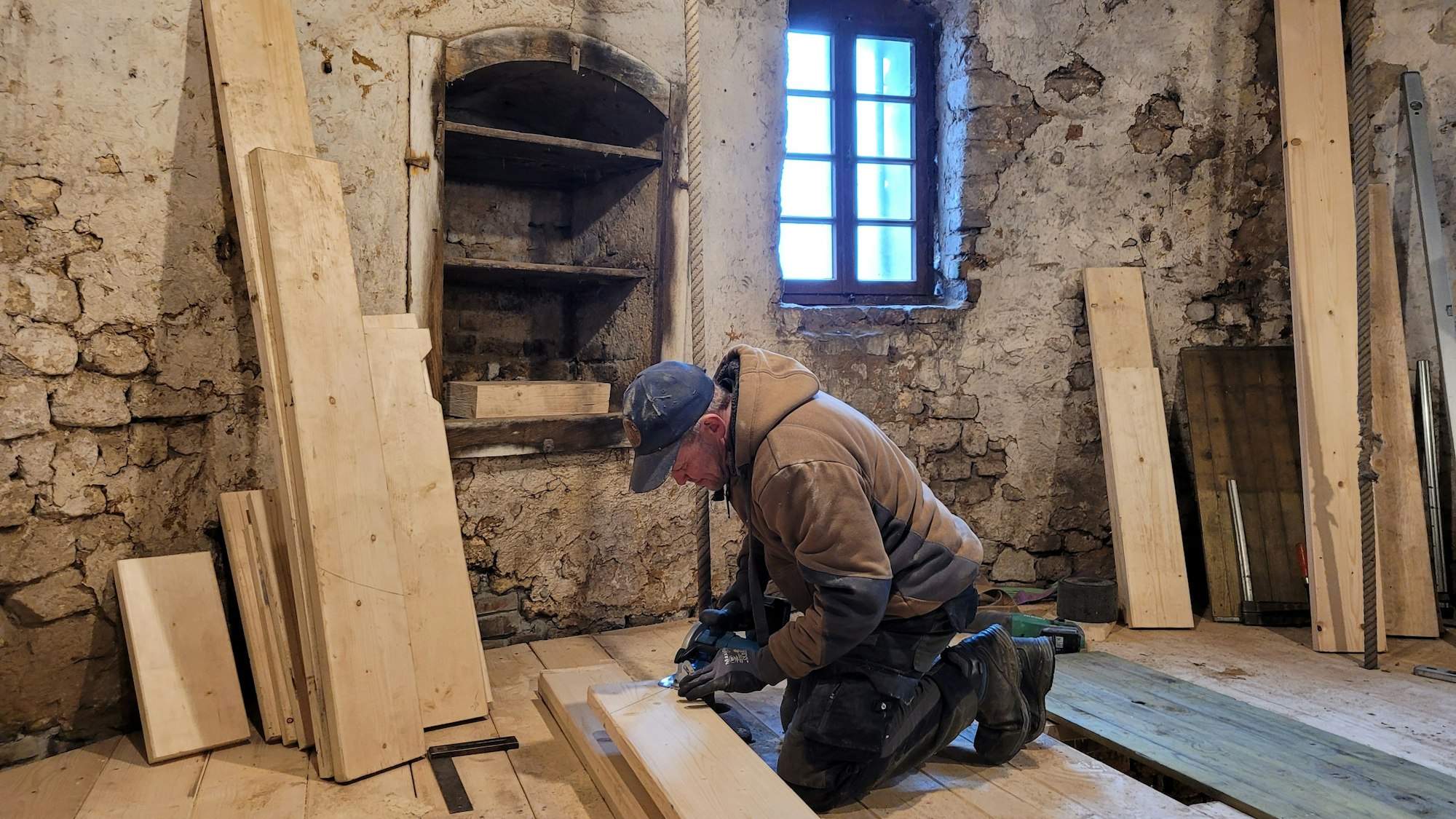 Ein Arbeiter kniet auf dem Boden, er schneidet ein Fußbodenbrett zurecht. An den Wänden lehnen unterschiedlich große Bretter.