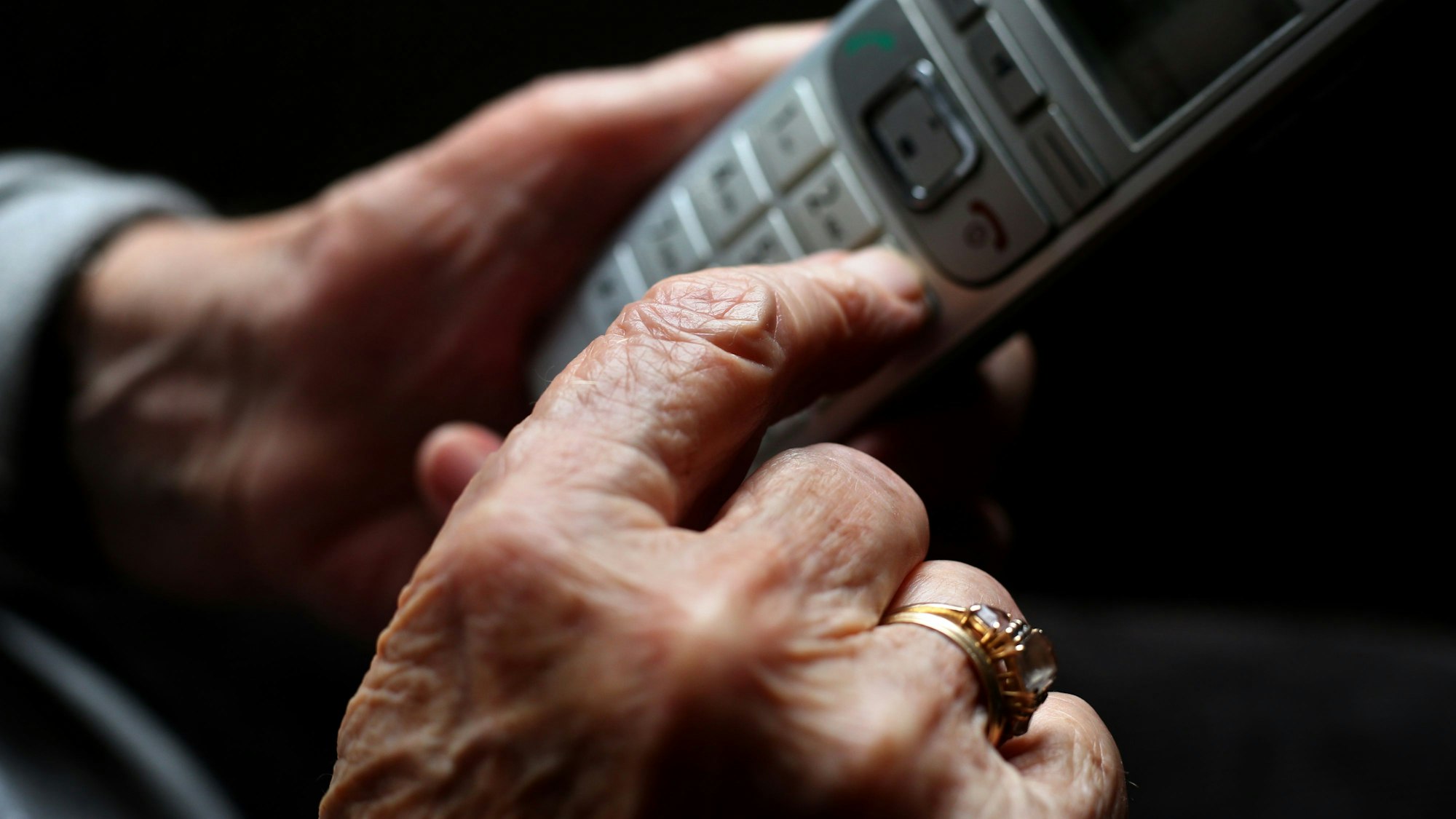 Eine ältere Frau tippt auf einem schnurlosen Festnetztelefon. An ihrer Hand ist ein großer Ring.