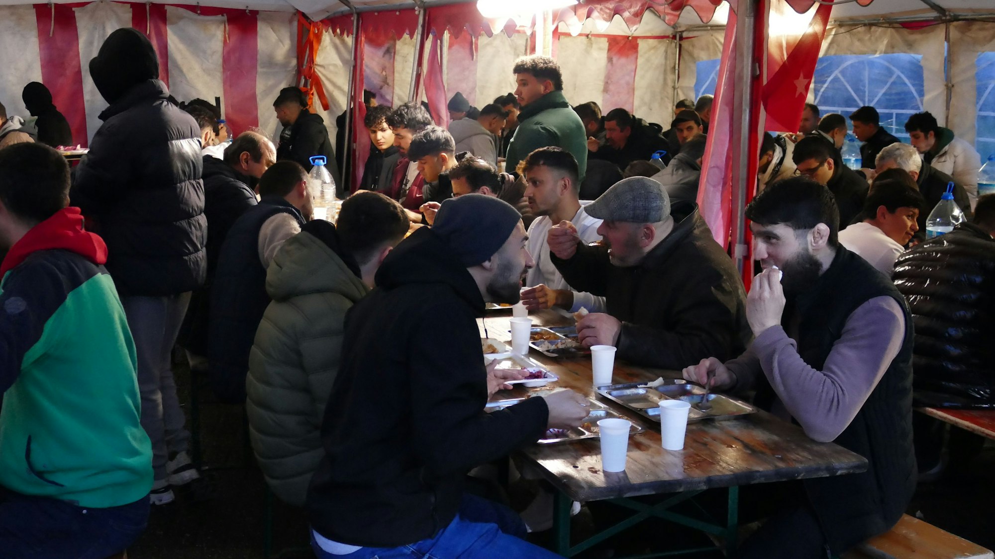 Männer sitzen an Biertischen in einem Zelt, sie essen.