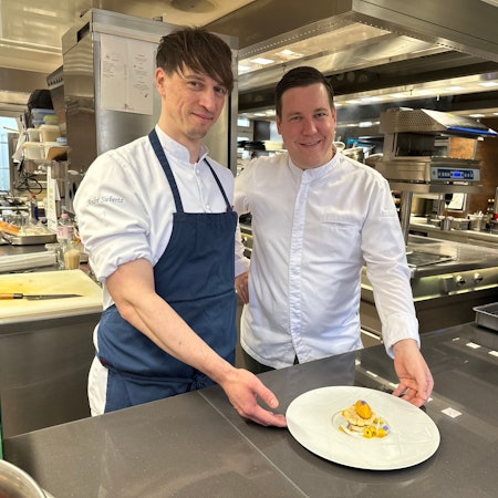 Zwei Männer stehen in einer Restaurant-Küche und zeigen einen elegant angerichtetes Gericht auf einem weißen Teller.