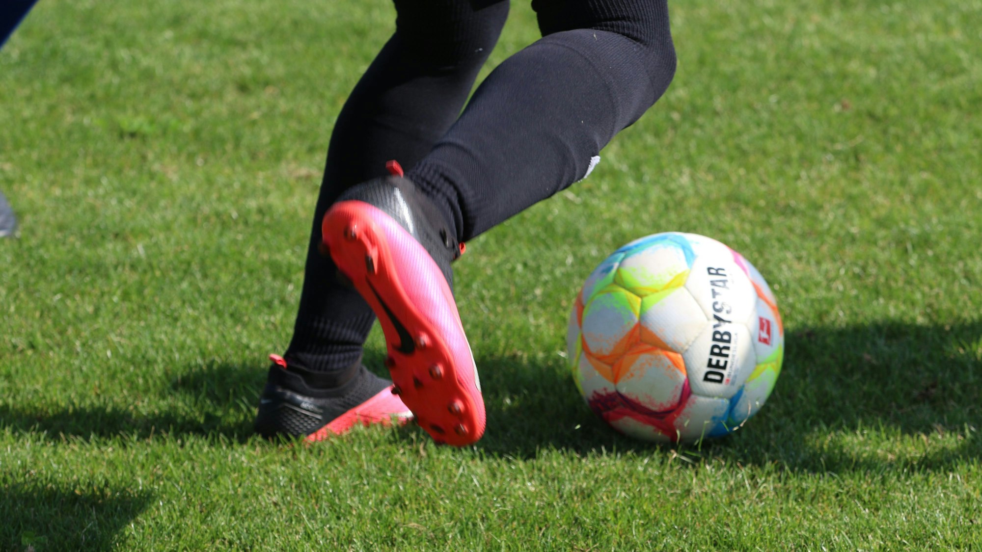 Die Füße eines Fußballspielers in pink-schwarzen Schuhen sind zu sehen, kurz bevor er einen Ball schießt.