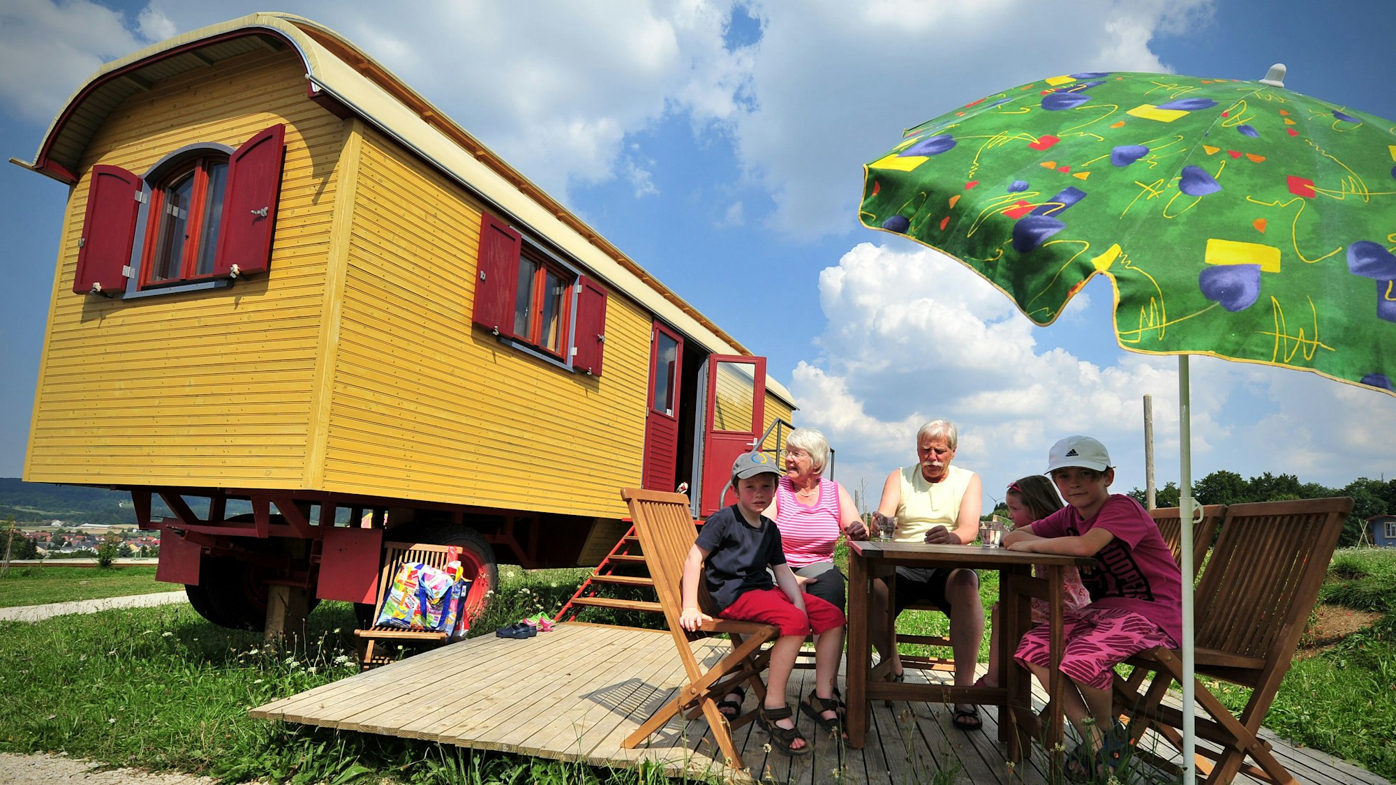 Ein gelber Zirkuswagen, davor sitzen Großeltern mit drei Kindern unter einem Sonnenschirm am Tisch.