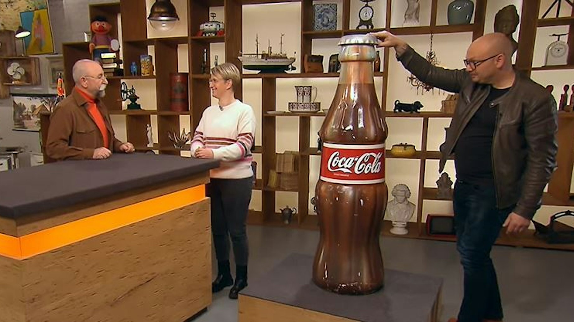 Experte Sven Deutschmanek (r.) nimmt die riesige Cola-Flaschen bei Bares für Rares in Augenschein, während Horst Lichter (l.) mit Verkäuferin Katrin Stegner (M.) spricht.
