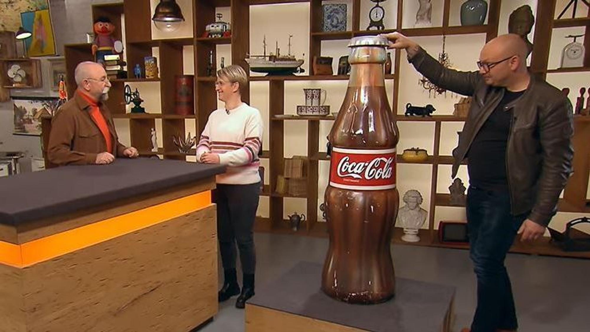 Experte Sven Deutschmanek (r.) nimmt die riesige Cola-Flaschen bei Bares für Rares in Augenschein, während Horst Lichter (l.) mit Verkäuferin Katrin Stegner (M.) spricht.