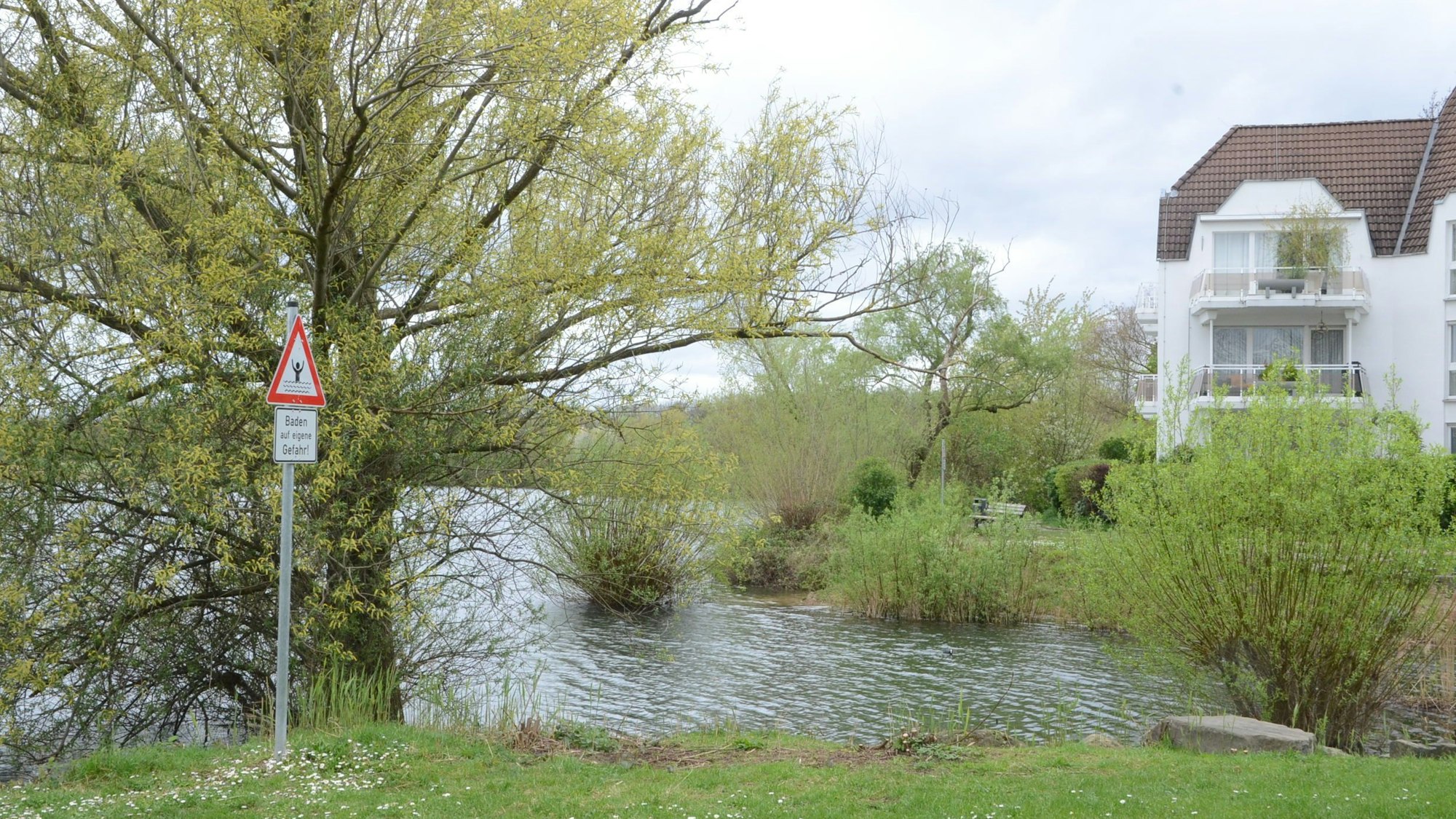 Bäume und Büsche an einem Seeufer. Ein Schild erklärt: Baden auf eigene Gefahr.