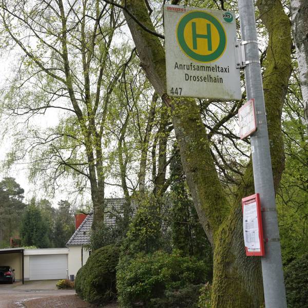 Man sieht das Haltestellenschild, im Hintergrund sind Bäume und Wohnhäuser.
