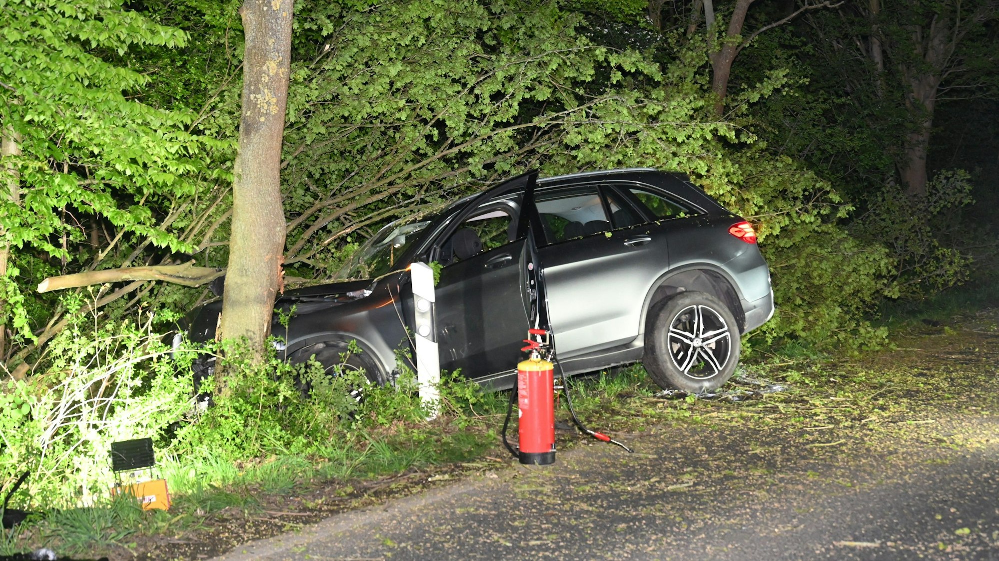 Ein Mercedes liegt in einem Graben. Er ist gegen einen Baum gefahren, ein abgebrochener Ast liegt vor dem Wagen. Neben der Fahrertür steht ein Feuerlöscher.