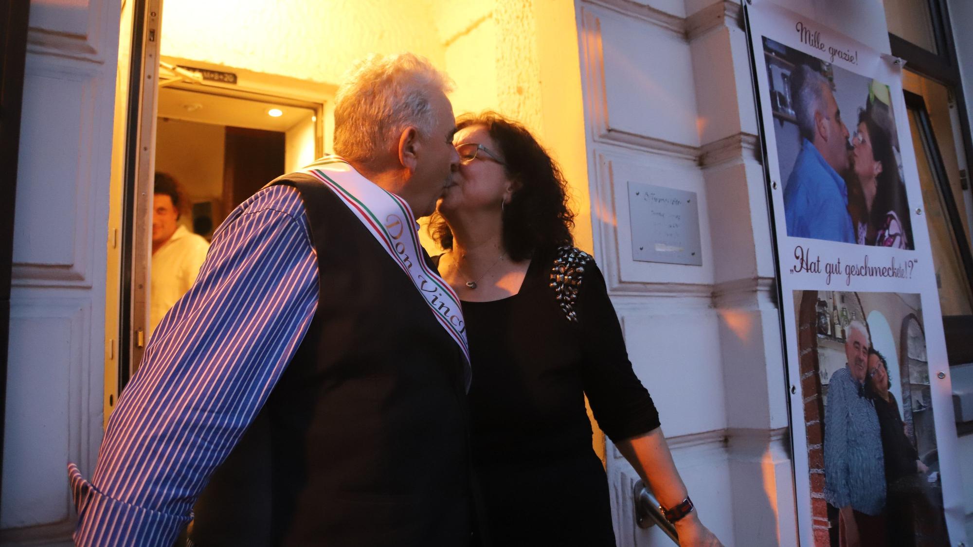Ein grauhaariger Mann und eine dunkelhaarige Frau stehen in einem Hauseingang, er trägt eine Schärpe, sie ein schwarzes Kleid. Sie küssen sich.