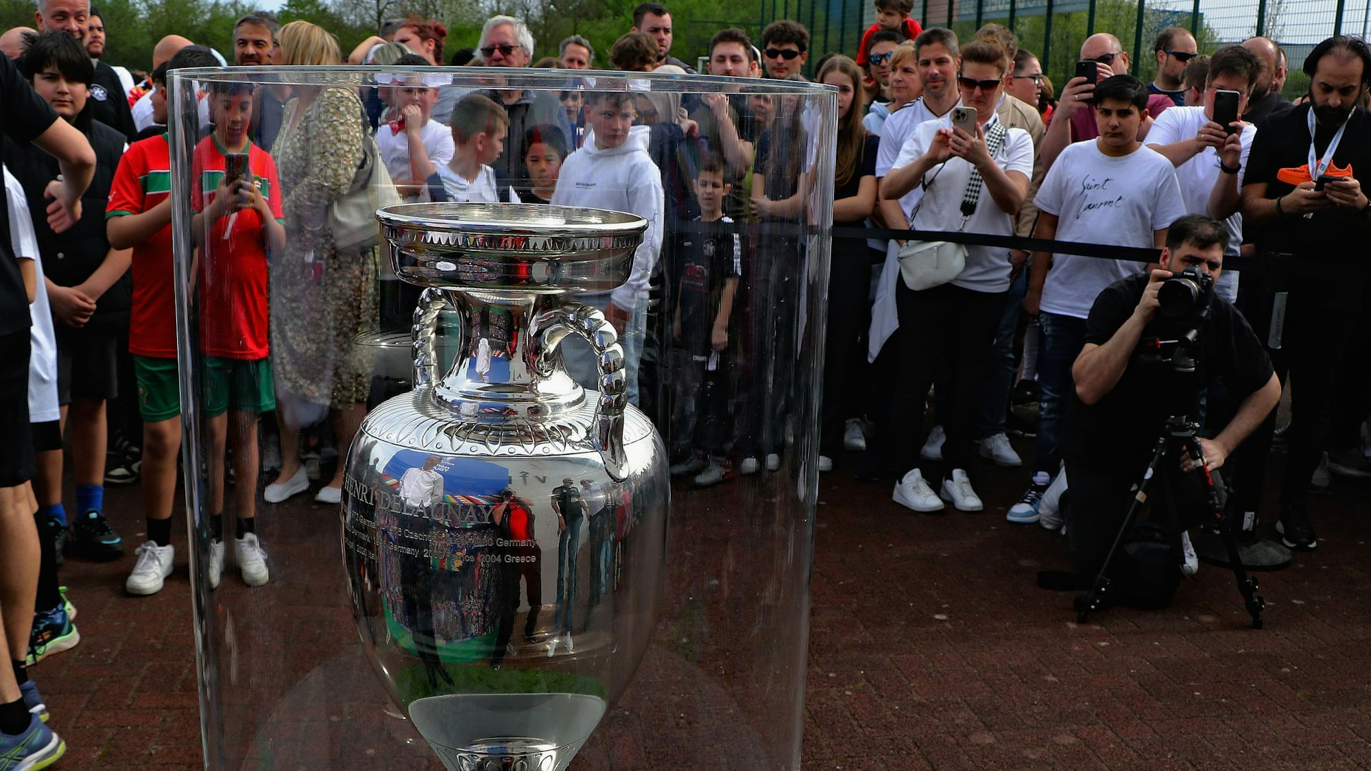 Zu sehen ist der EM-Pokal in einer Vitrine, im Hintergrund stehen viele Menschen, die Fotos machen.